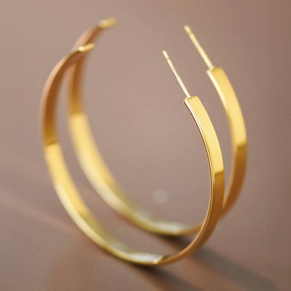 A pair of polished gold hoop earrings with a modern, slightly tapered barbell-like design on the front of each hoop, shown on a neutral warm brown background.