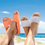 A pair of feet wearing orange and white slides, with the beach and blue sky in the background.