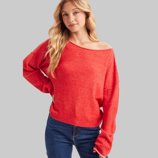 A woman wearing a bright red off-the-shoulder knit sweater and dark blue jeans, photographed against a clean light gray studio background.