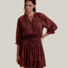 A woman modeling a short, loose-fitting dress with a dark red and brown paisley pattern, featuring a keyhole tie neckline, 3/4 puff sleeves, and a smocked waistband, set against a solid beige background.