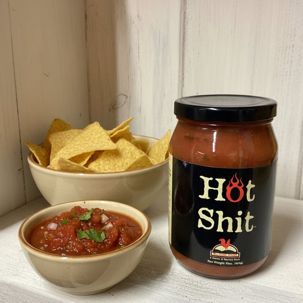 Jar of salsa labeled 'Hot Shit' next to a bowl of chips and salsa on a wooden surface.