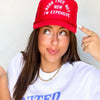Person wearing a red cap with text and a white shirt, pointing at the cap.
