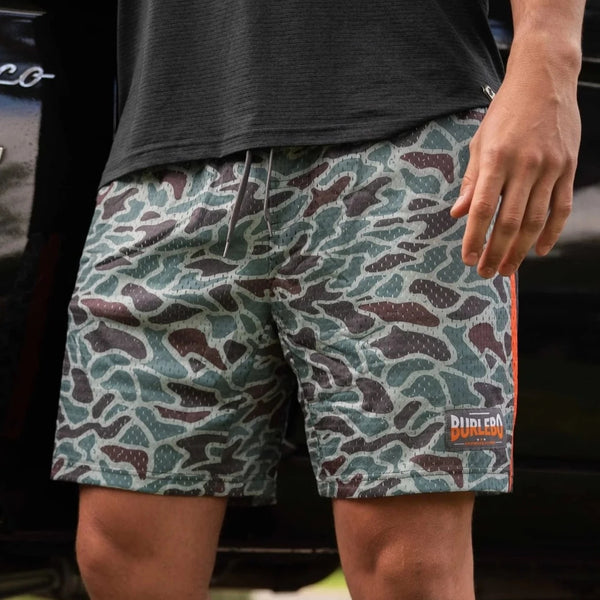 Person wearing camouflage shorts with a brand logo, standing in front of a blurred background.