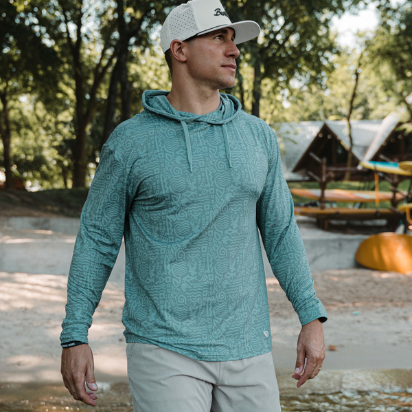 Man wearing a teal patterned hoodie and light gray cap outdoors with trees and a wooden structure in the background.