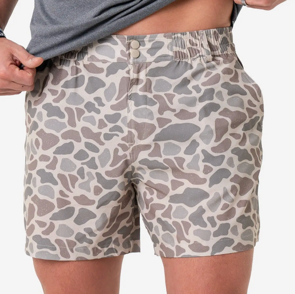 A close-up of a person wearing patterned shorts with a gray shirt partially pulled up. The shorts have an abstract organic spot pattern in beige, gray, and taupe tones.