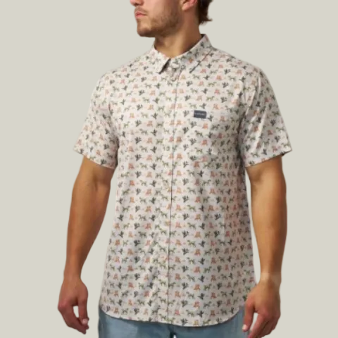 A man wearing a short-sleeve button-up shirt with a dense, small-scale multicolor pattern of birds and foliage on a light beige background. The shirt features a small dark logo patch on the left chest.