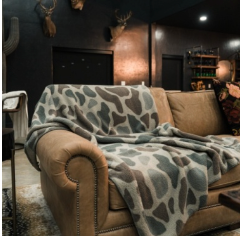 A cozy brown leather sofa in a dimly lit room, draped with a plush throw blanket featuring a gray, beige, and white organic animal-like pattern.
