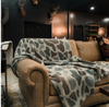 A cozy brown leather sofa in a dimly lit room, draped with a plush throw blanket featuring a gray, beige, and white organic animal-like pattern.