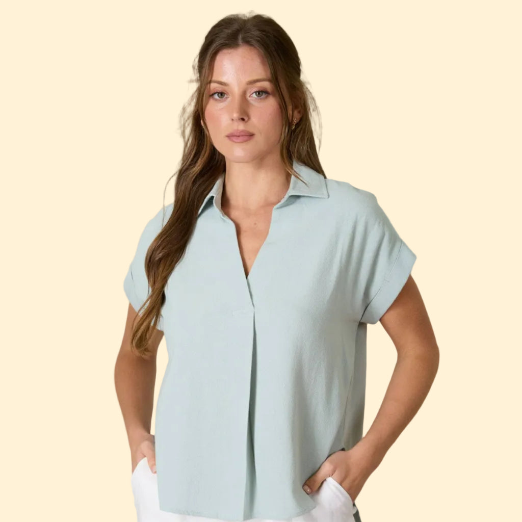 Woman wearing a light blue shirt and white pants in a bright room with plants.