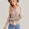A woman wearing a taupe long-sleeve henley top with lace sleeve cuffs, paired with blue denim shorts and a brown wide-brim hat, against a plain white studio background.