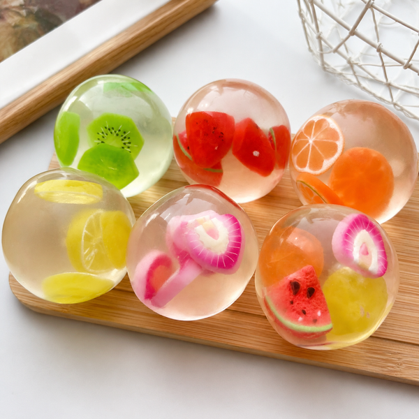 Six translucent gel spheres filled with fruit slices and pieces are arranged on a wooden board. The spheres contain colorful fruit designs including kiwi, watermelon, orange, and pink radish slices.