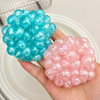 A hand holds two iridescent bubble-like gel balls, one turquoise blue and one soft pink, against a light neutral background with a white plate behind them.