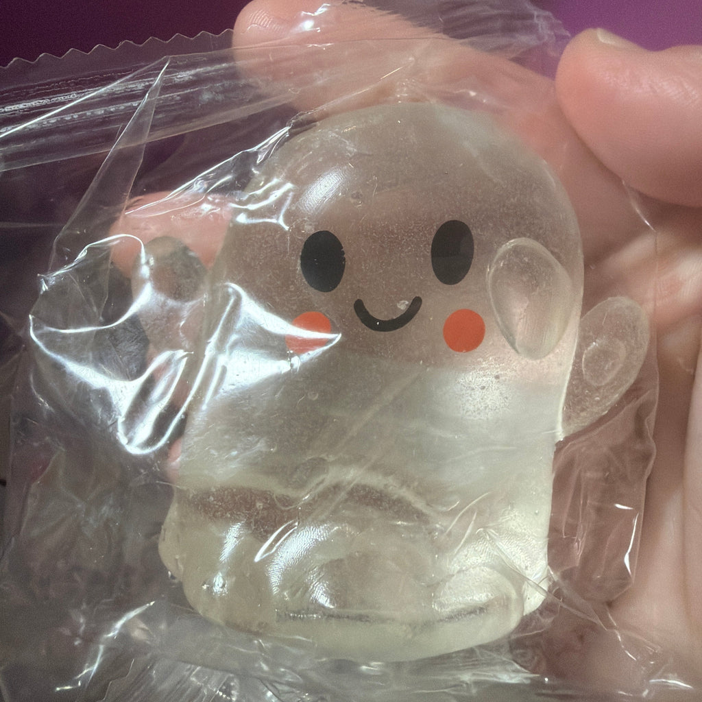 A small translucent ghost-shaped toy or figurine with a simple black face and orange cheeks, still inside clear plastic packaging and being held by a hand.