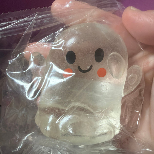 A small translucent ghost-shaped toy or figurine with a simple black face and orange cheeks, still inside clear plastic packaging and being held by a hand.