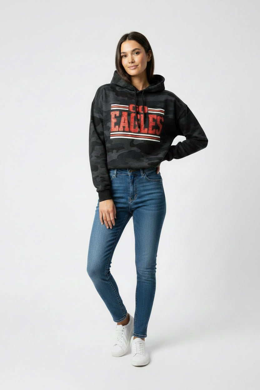 A woman modeling a black camouflage hoodie with a red and white 'GO EAGLES' graphic on the chest, paired with blue skinny jeans and white sneakers, against a light gray studio background