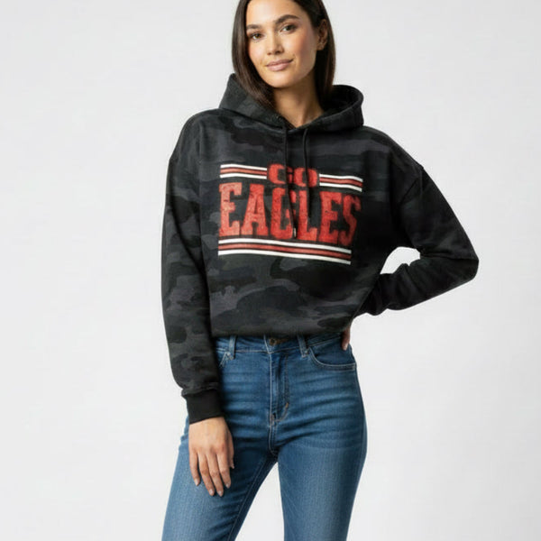 A woman modeling a black camouflage hoodie with a red and white 'GO EAGLES' graphic on the chest, paired with blue skinny jeans and white sneakers, against a light gray studio background