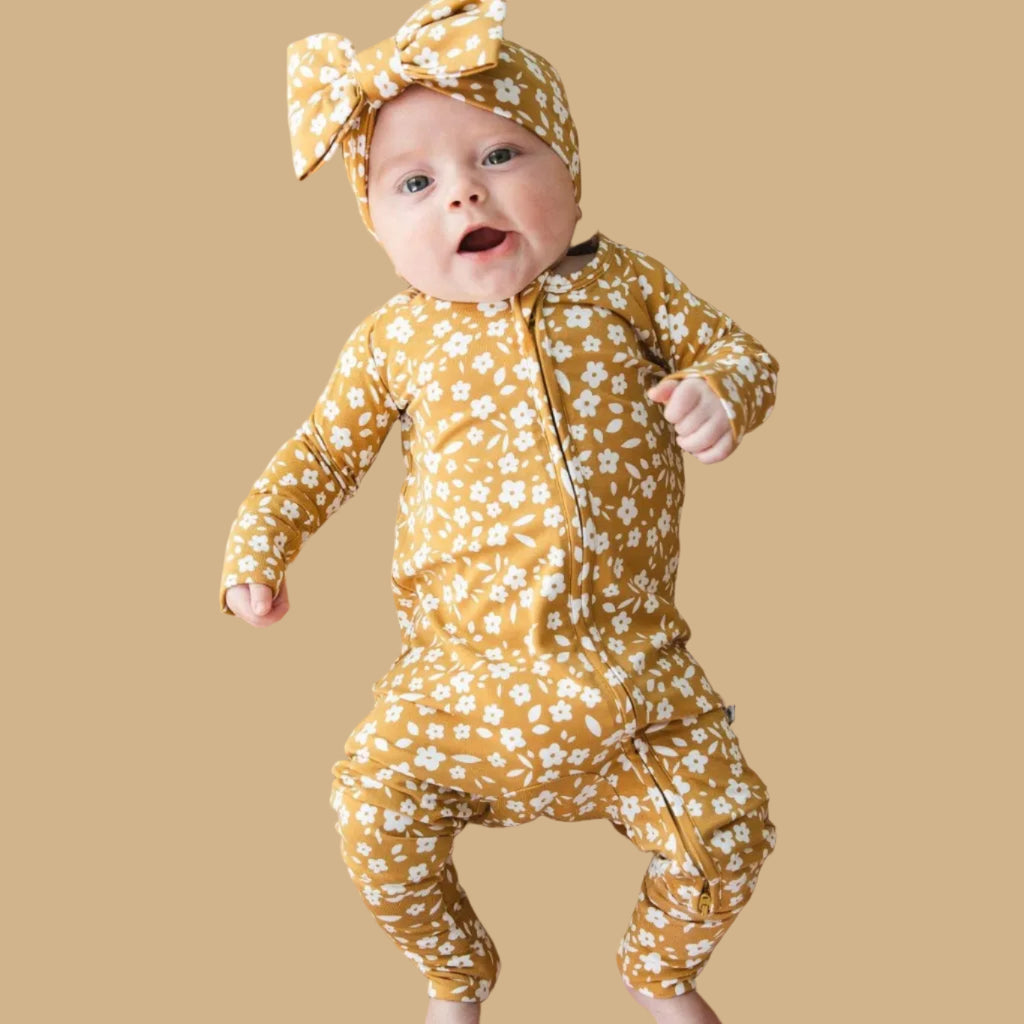 Baby in a yellow floral outfit and headband lying on a textured mustard yellow blanket.