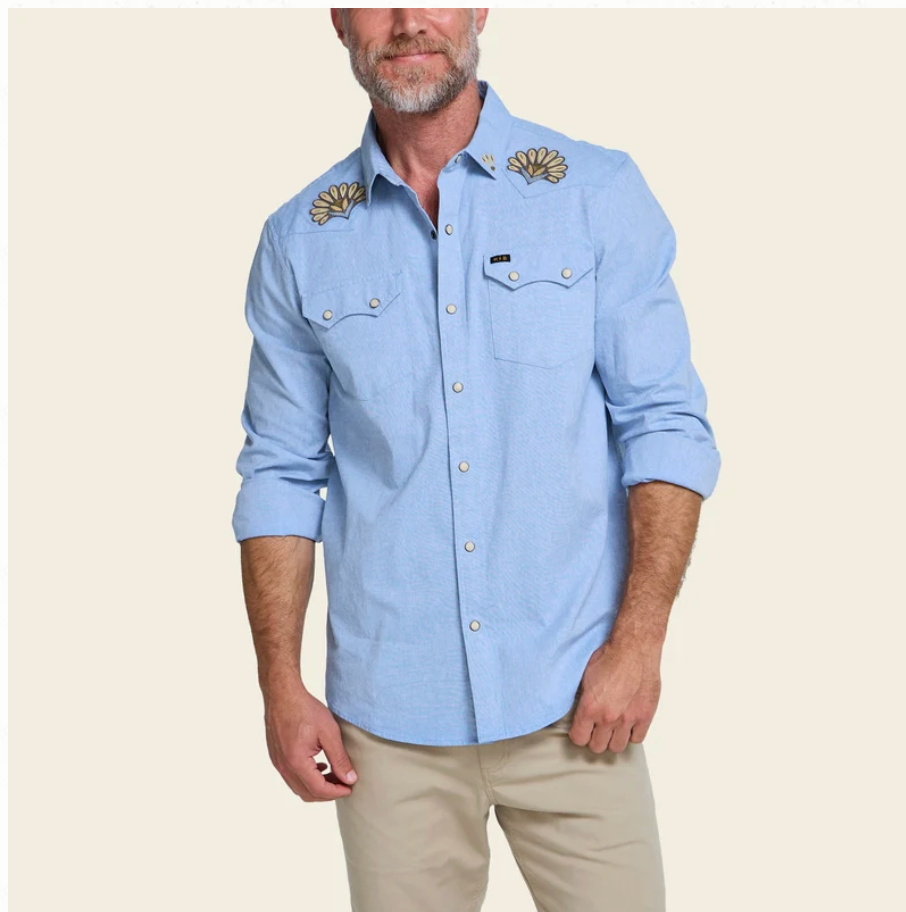 A man wearing a light blue long-sleeve western-style button-up shirt with embroidered floral details on the shoulders and beige pants, standing against a plain light beige background.