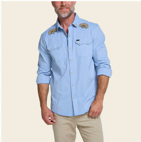 A man wearing a light blue long-sleeve western-style button-up shirt with embroidered floral details on the shoulders and beige pants, standing against a plain light beige background.