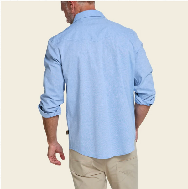 A man shown from the back wearing a light blue long-sleeve button-down shirt with rolled-up sleeves and khaki pants, set against a solid light beige background.