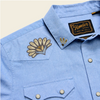 Close-up of a light blue western-style button-down shirt with a pointed collar, embroidered floral patch on the chest, and a visible Flomler Bros. neck label.