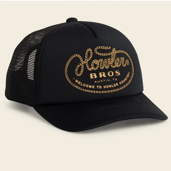 A black trucker hat with a curved brim and black mesh back panels, featuring a gold embroidered logo on the front that reads 'Howler Bros. Austin, TX' with 'Welcome to Howler County' below, set against a light beige background.