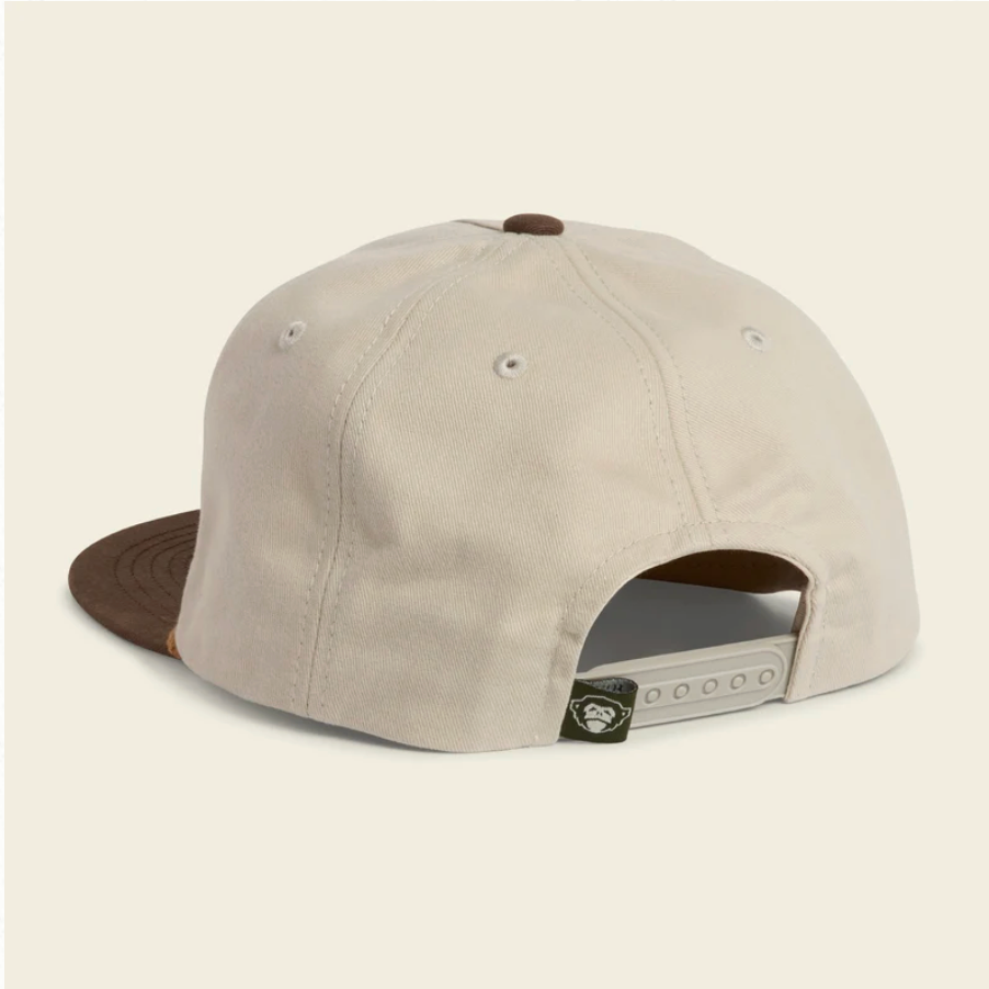 A light beige snapback cap shown from the back, featuring a brown flat brim, brown button on top, and a white plastic adjustment slider with a small black brand patch.