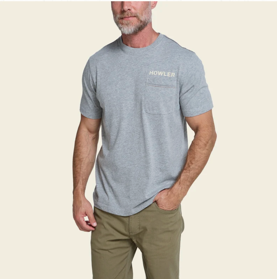 A man wearing a heather gray short-sleeve t-shirt with a chest pocket and 'HOWLER' printed on the left chest, paired with olive green pants, against a solid light beige background.