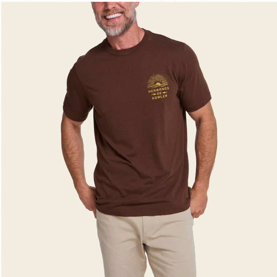 A man wearing a brown crewneck t-shirt with a small gold graphic on the left chest that reads 'Hermanos OofHowler, paired with beige trousers, against a solid light beige background.