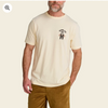 A man wearing a cream-colored short-sleeve t-shirt with a small black and brown graphic on the left chest that reads 'HONOR US' above a bear illustration. The background is a solid light cream color.
