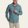 A man wearing a muted blue-green long-sleeve button-up shirt with two chest flap pockets, paired with khaki pants, against a solid light beige background.