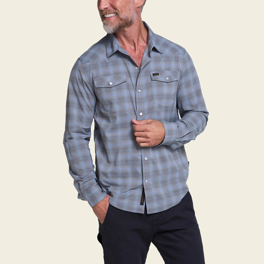A man wearing a long-sleeve plaid button-up shirt in shades of blue and gray, paired with dark pants, against a solid light beige background.