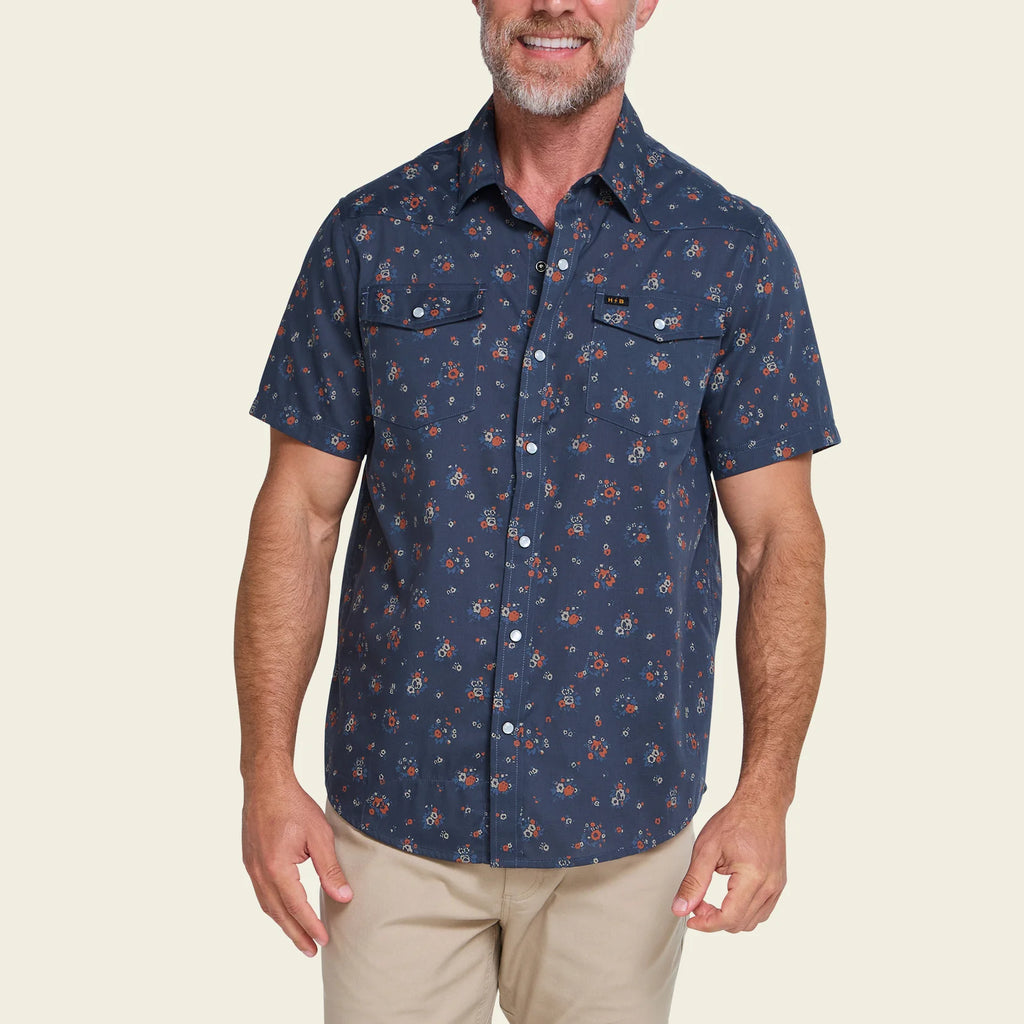 A man wearing a short-sleeve button-up shirt with a navy blue base and a small red and orange floral pattern, paired with khaki pants, against a solid light beige background.