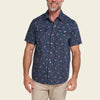 A man wearing a short-sleeve button-up shirt with a navy blue base and a small red and orange floral pattern, paired with khaki pants, against a solid light beige background.