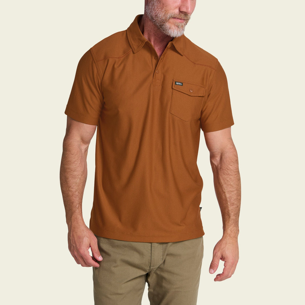 A man wearing a rust-brown short-sleeve polo shirt with a chest pocket, paired with khaki pants, shown against a light beige background.