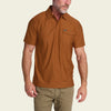 A man wearing a rust-brown short-sleeve polo shirt with a chest pocket, paired with khaki pants, shown against a light beige background.