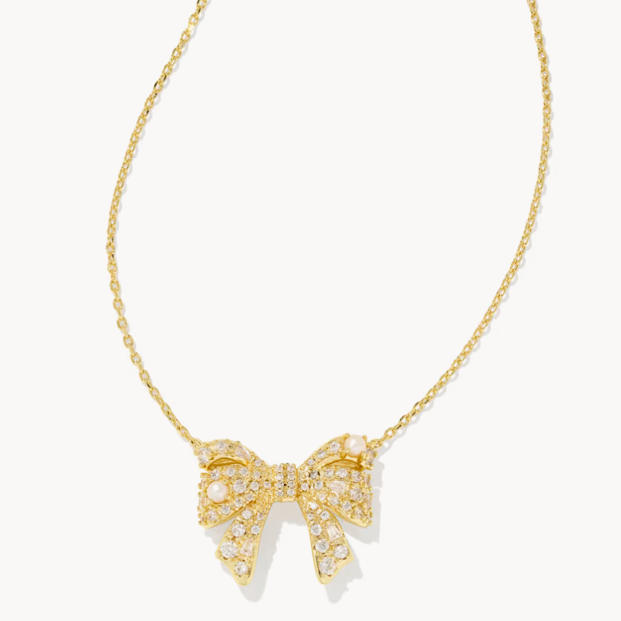 A gold chain necklace featuring a bow-shaped pendant paved with small clear stones and accented with white pearls, shown against a light off-white background.