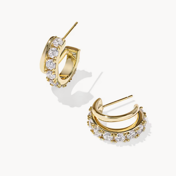 A pair of gold huggie hoop earrings adorned with clear pavé gemstones, displayed on a clean off-white background.