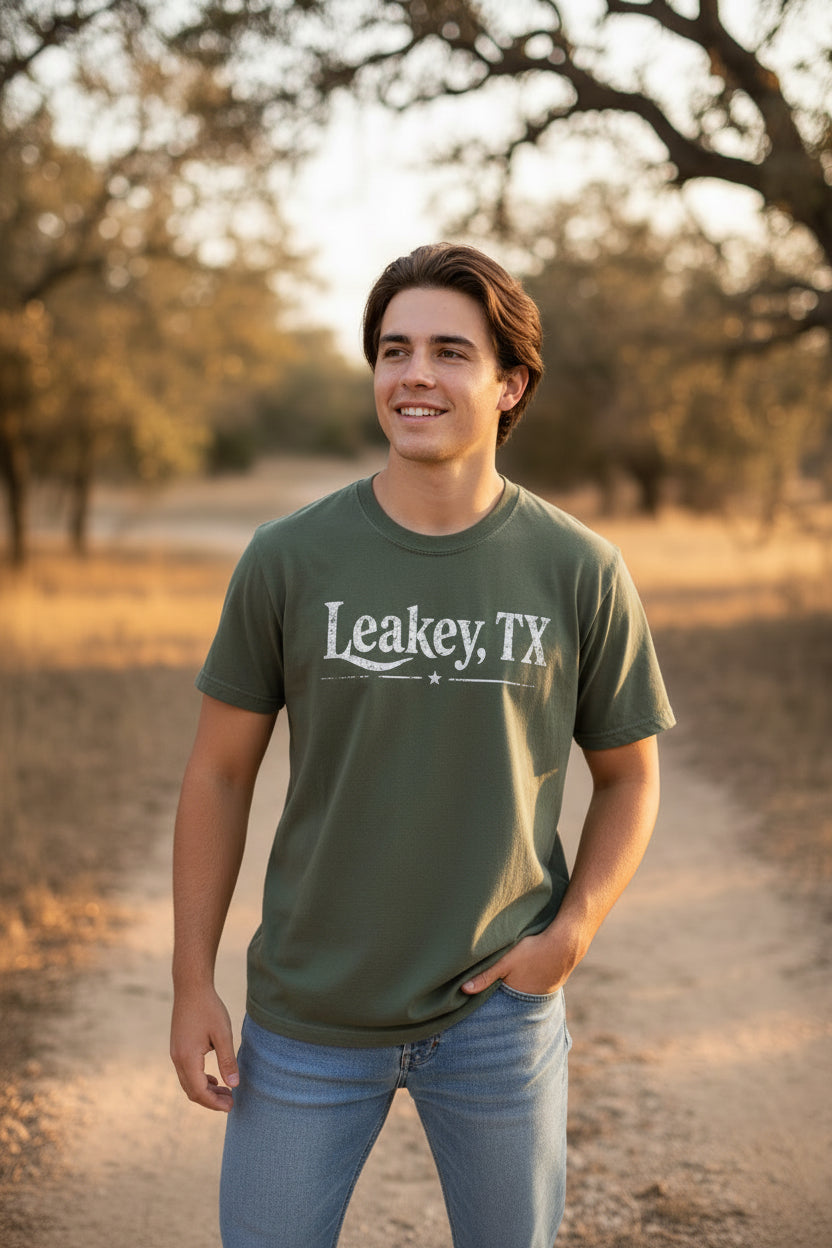 An olive green t-shirt on a model, featuring the text 'Leakey, TX' in a distressed white font across the chest.