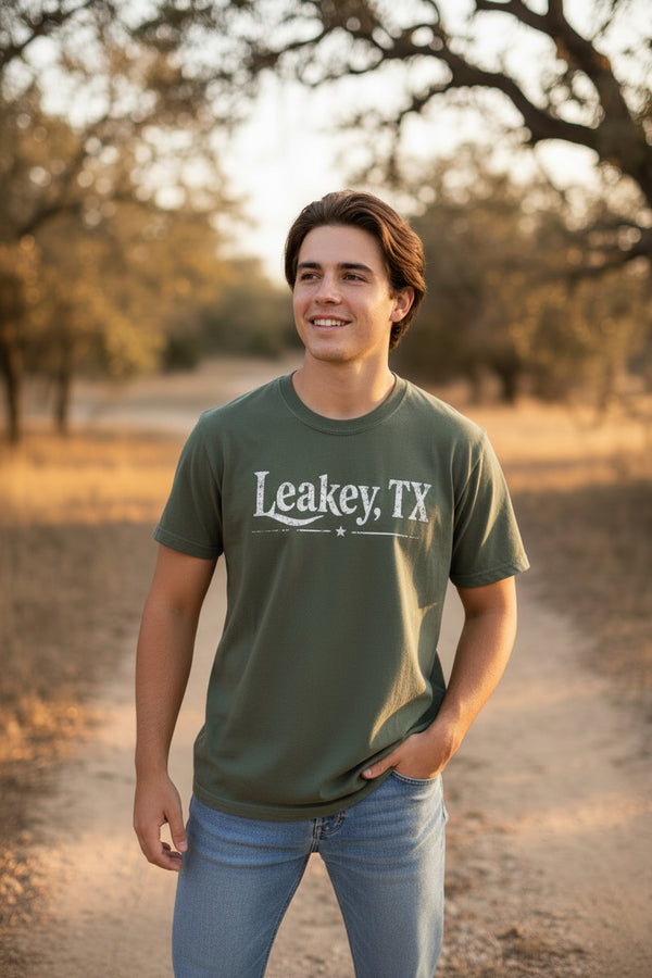 An olive green t-shirt on a model, featuring the text 'Leakey, TX' in a distressed white font across the chest.