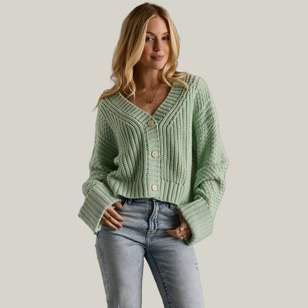 Woman wearing a light green knitted cardigan and blue jeans against a gray background