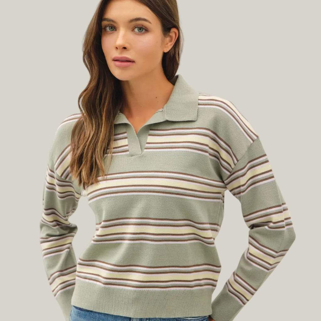 A woman wearing a sage green long-sleeve polo sweater with horizontal stripes in beige, brown, and muted pink, paired with blue jeans, against a plain light gray studio background.