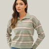 A woman wearing a sage green long-sleeve polo sweater with horizontal stripes in beige, brown, and muted pink, paired with blue jeans, against a plain light gray studio background.