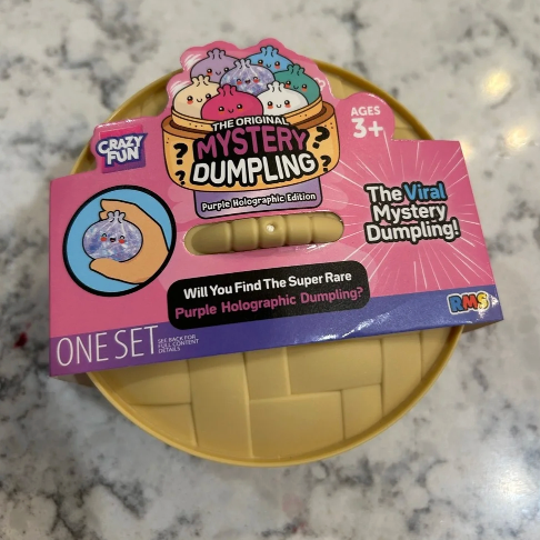 Crazy Fun The Original Mystery Dumpling toy set in its retail packaging, featuring a pink and purple box with a clear window showing the yellow dumpling inside, placed on a yellow circular tray over a marble countertop.