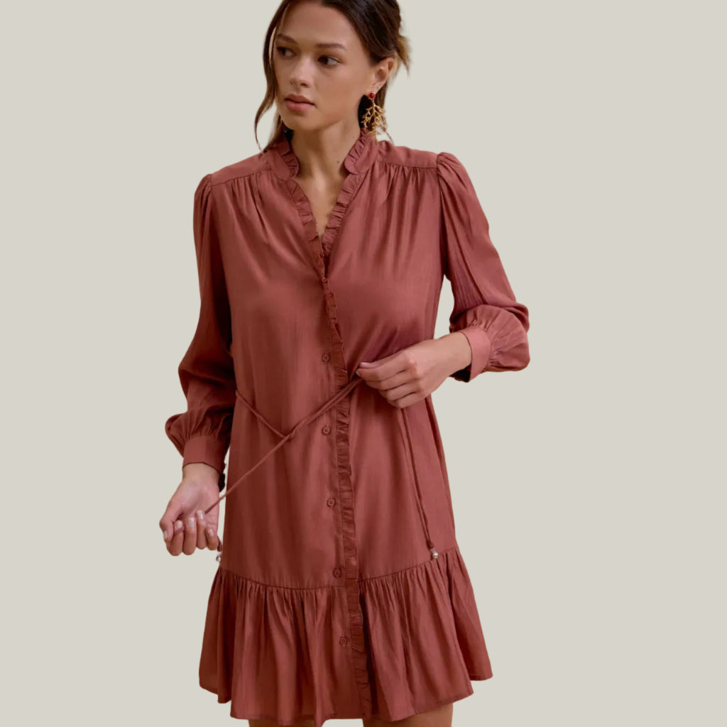 A woman wearing a dusty rose long-sleeve mini dress with a ruffled collar and waist tie, posed against a solid beige background.