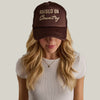 Woman wearing a brown cap with 'Raised on Country' text against a gray background