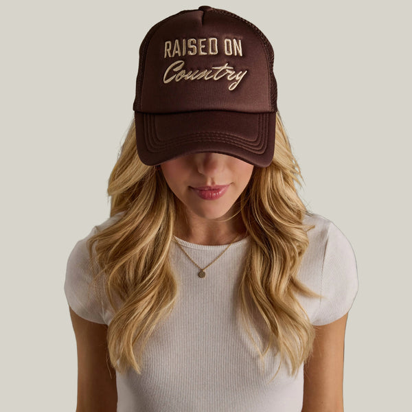 Woman wearing a brown cap with 'Raised on Country' text against a gray background