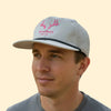 Man wearing a cap with pink antlers and 'River Boyz' text on a beige background