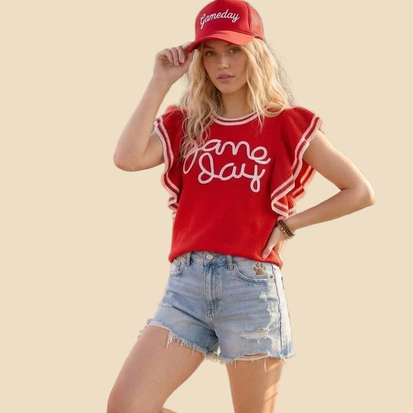 A woman stands wearing a red graphic t-sweater with 'Game Day' in white script, light blue denim shorts, a red baseball cap, and white sneakers. 