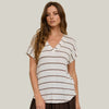 A woman wearing a white short-sleeve V-neck top with thin brown horizontal stripes, paired with dark brown wide-leg pants, standing against a beige background